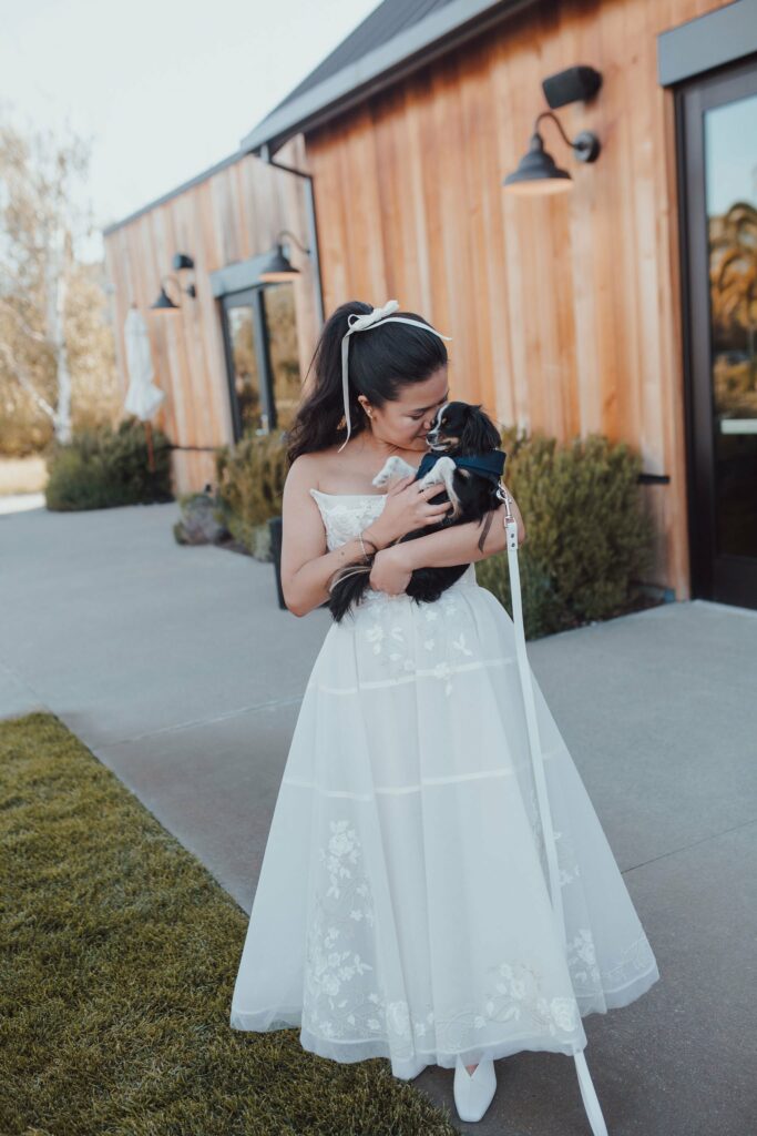 A woman in a white dress holds a black dog on a leash outside a wooden building.