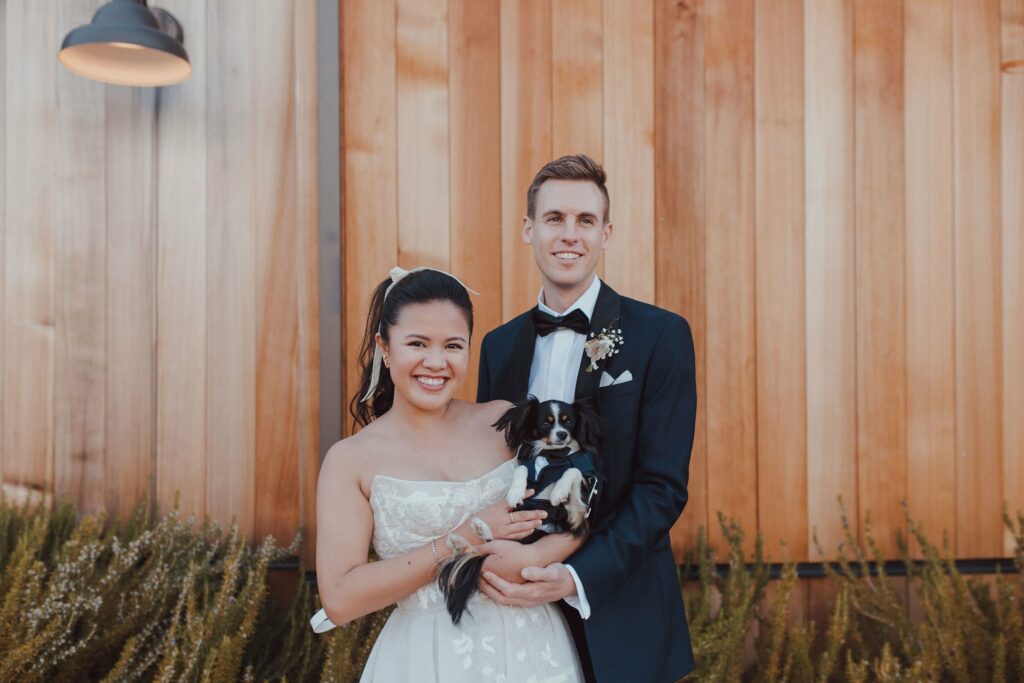 A couple in formal wedding attire stands outside in front of a wooden wall, smiling and holding a small black and white dog.