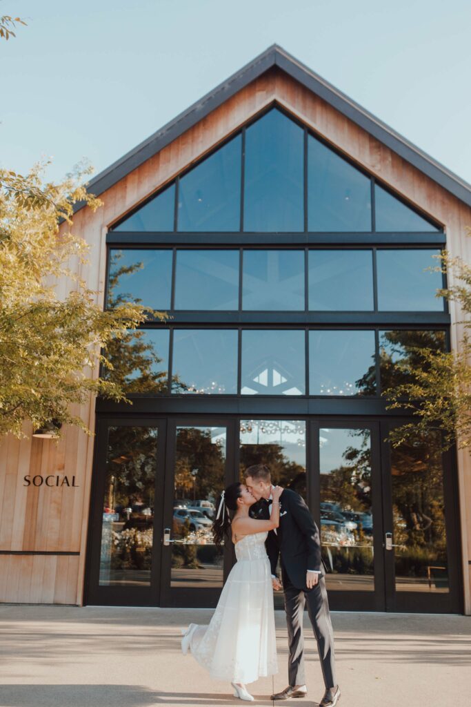 A couple in wedding attire shares a kiss in front of a modern building with large glass windows and a sign that reads "SOCIAL.