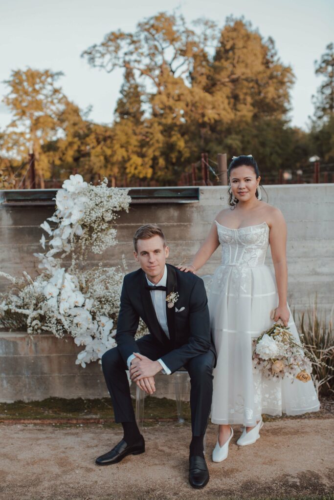 A bride and groom pose outdoors in wedding attire. The groom sits on a bench while the bride stands next to him holding a bouquet. A floral arrangement decorates the background.