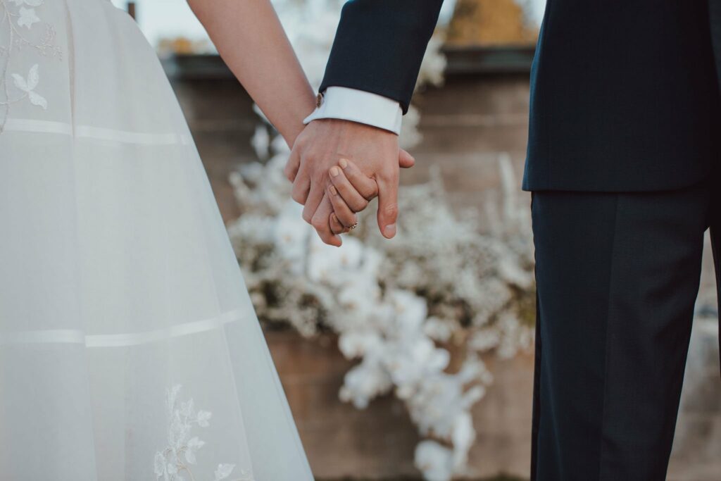 A bride and groom hold hands, both wearing formal attire. The bride's dress is white with floral embroidery and the groom's suit is dark. Blurred floral decorations are visible in the background.