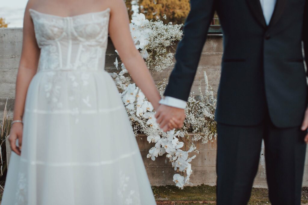 A bride in a strapless white wedding dress and a groom in a dark suit stand holding hands, with a floral arrangement in the background.