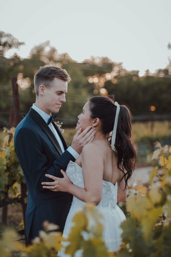 A couple dressed in wedding attire shares an intimate moment in an outdoor setting with greenery and a soft-focus background.