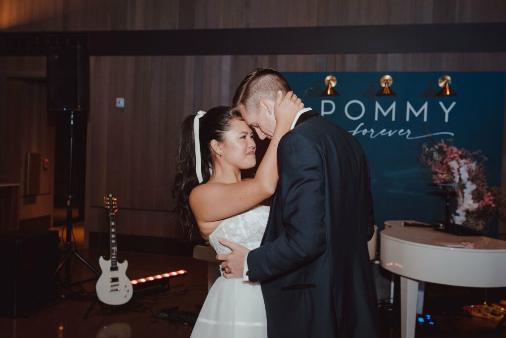 A couple in formal attire, with the woman in a white dress and the man in a dark suit, share a moment while dancing in front of a backdrop with the words "POMMY forever.