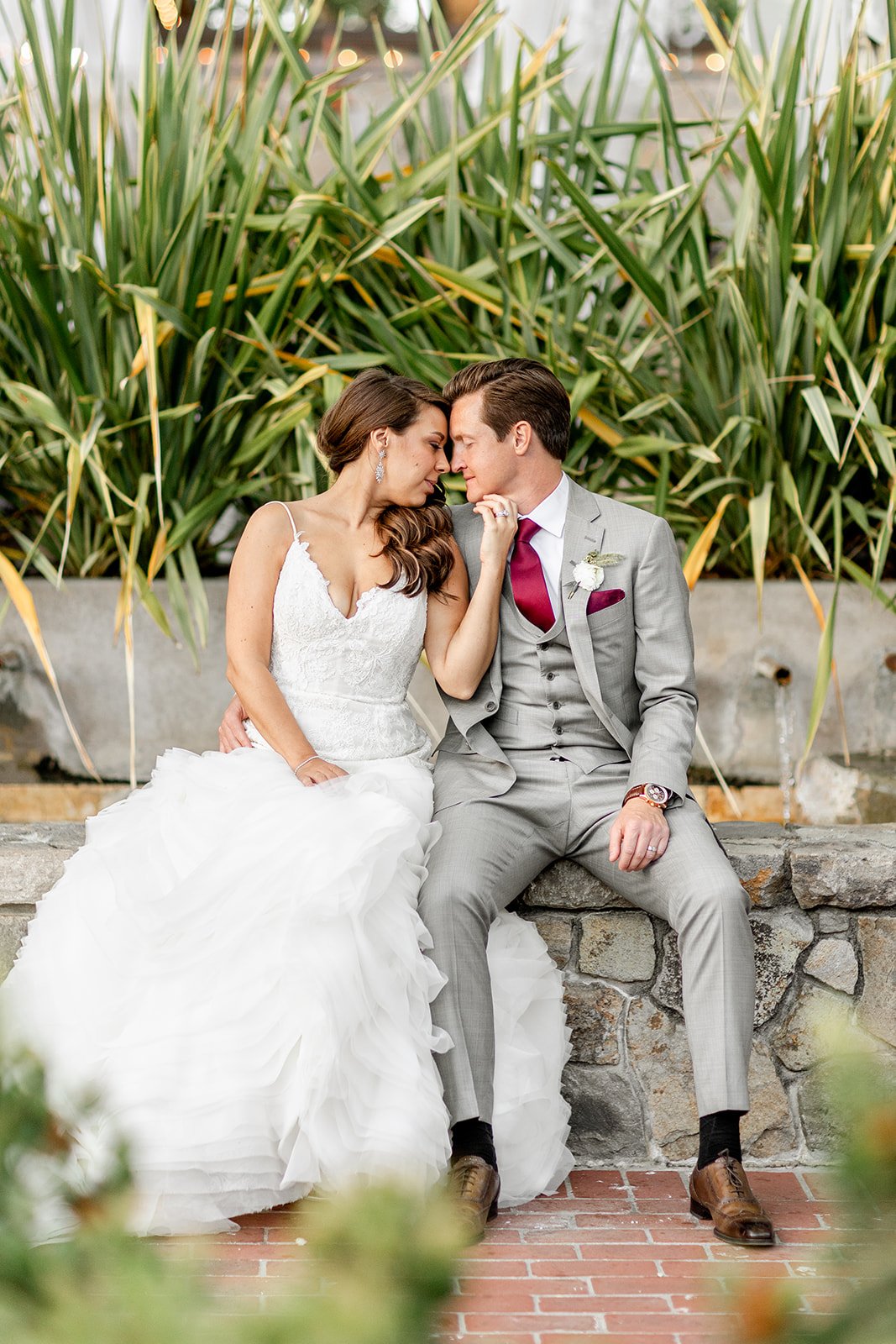 A bride in a white dress and groom in a gray suit with a red tie are sitting together on a stone ledge, leaning in and touching foreheads, surrounded by green plants.