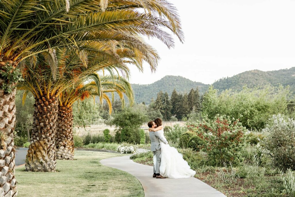 A couple in wedding attire embraces on a path with lush greenery and mountains in the background, flanked by palm trees.