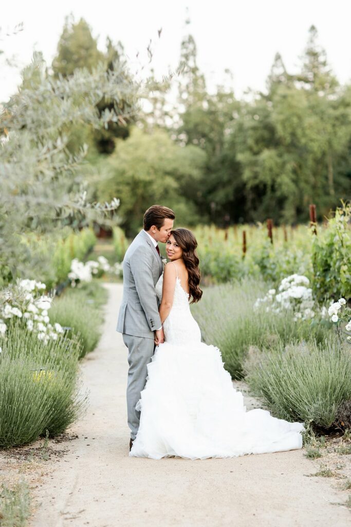 A bride and groom stand closely together on a path in a garden, surrounded by greenery and white flowers. She wears a white wedding dress and he is in a gray suit.