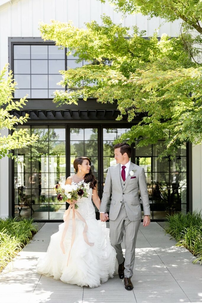 A couple walks hand-in-hand outside a building with large windows. The woman is in a white wedding dress holding a bouquet, and the man is in a light grey suit with a red tie.