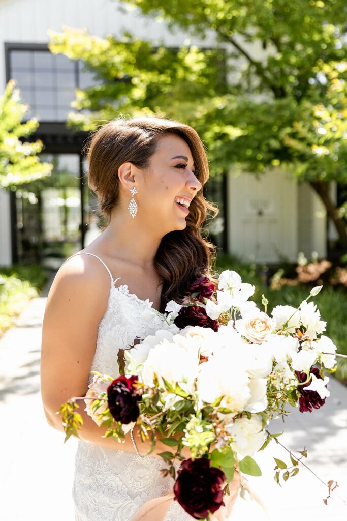 A bride smiles while holding a large bouquet of white and dark purple flowers. She is standing outdoors in front of a building with greenery in the background.