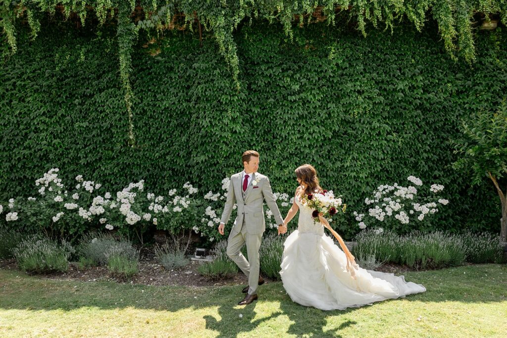 A couple in formal attire, with the bride in a white dress and the groom in a light suit, walk hand in hand in front of a green ivy-covered wall adorned with white flowers.