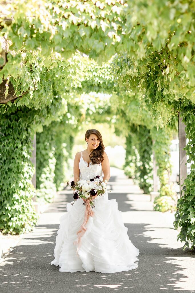 A bride in a white dress holds a bouquet while standing under a walkway covered with greenery.