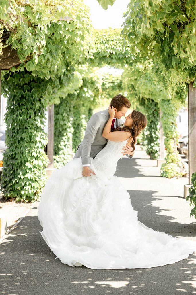 A groom in a grey suit and a bride in a white wedding dress share a romantic dip kiss under an archway covered with green ivy.