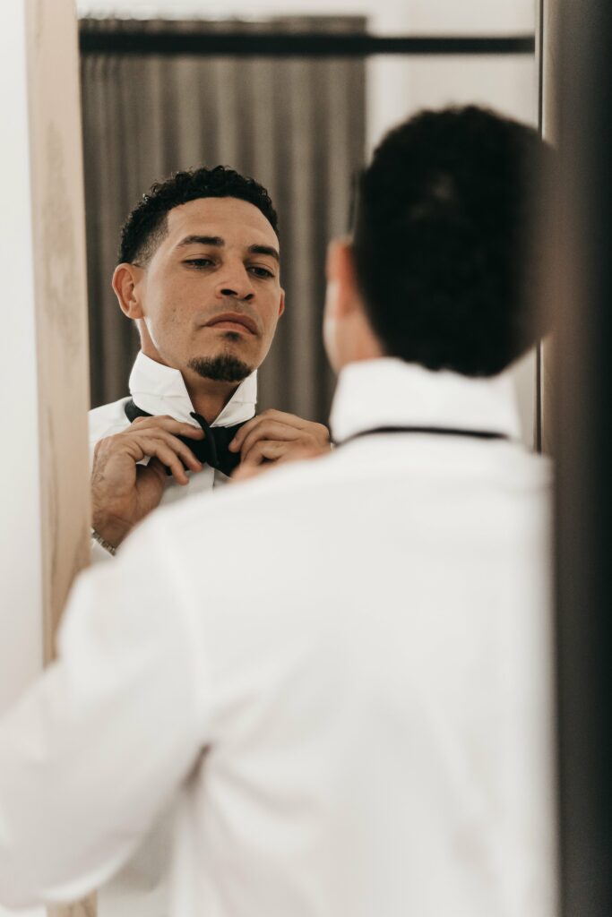 A man adjusts his black bow tie in front of a mirror, wearing a white dress shirt.