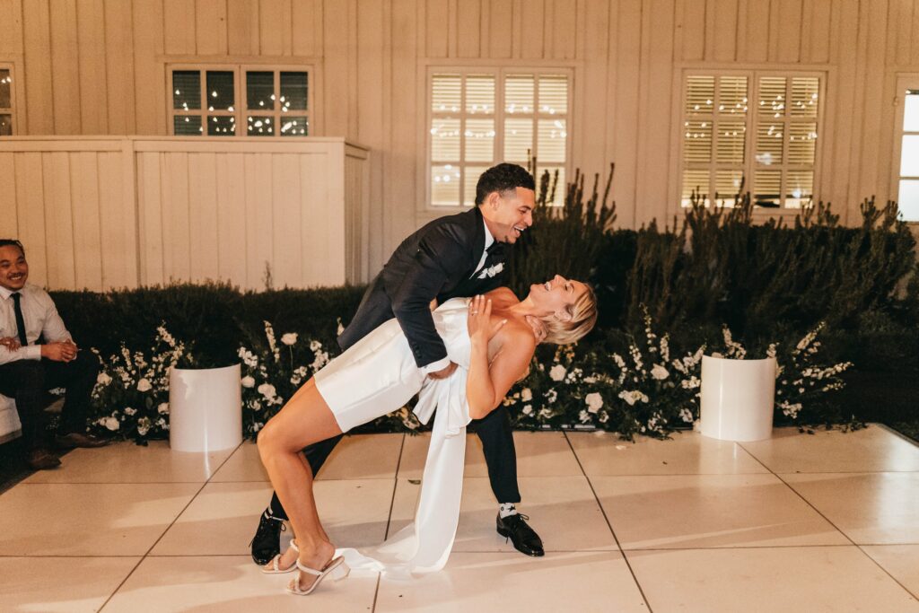 A couple dances closely, with one person in a black suit leaning back the other in a white dress on a white tiled floor.