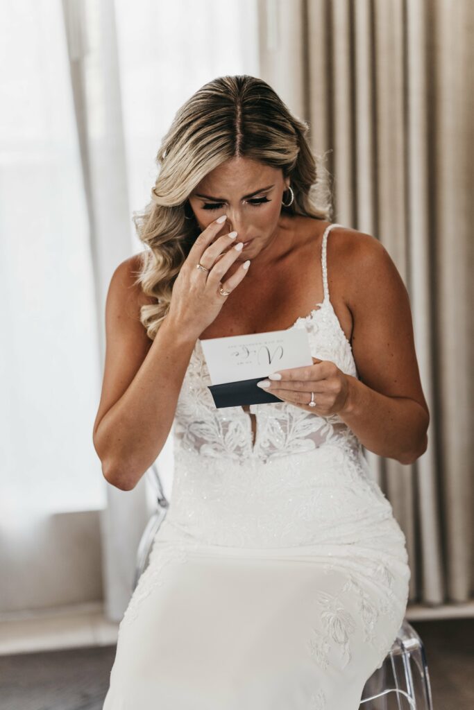 A woman in a white dress sits and reads a card, appearing emotional. She holds the card in one hand and touches her face with the other. She is in front of light-colored curtains.