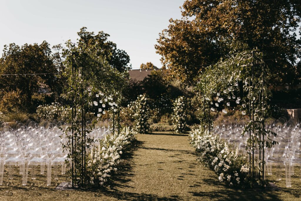Outdoor wedding ceremony setup with clear chairs arranged in rows, floral decorations on archways, and a green aisle at the center, surrounded by trees and greenery.