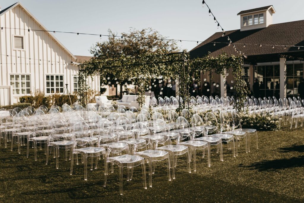 An outdoor wedding setup with rows of transparent chairs facing a floral-decorated arch. String lights are hung above, and white buildings are in the background.