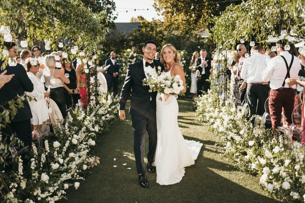 A smiling couple in wedding attire walks down an aisle decorated with white flowers and greenery, surrounded by clapping and cheering guests outdoors on a sunny day.