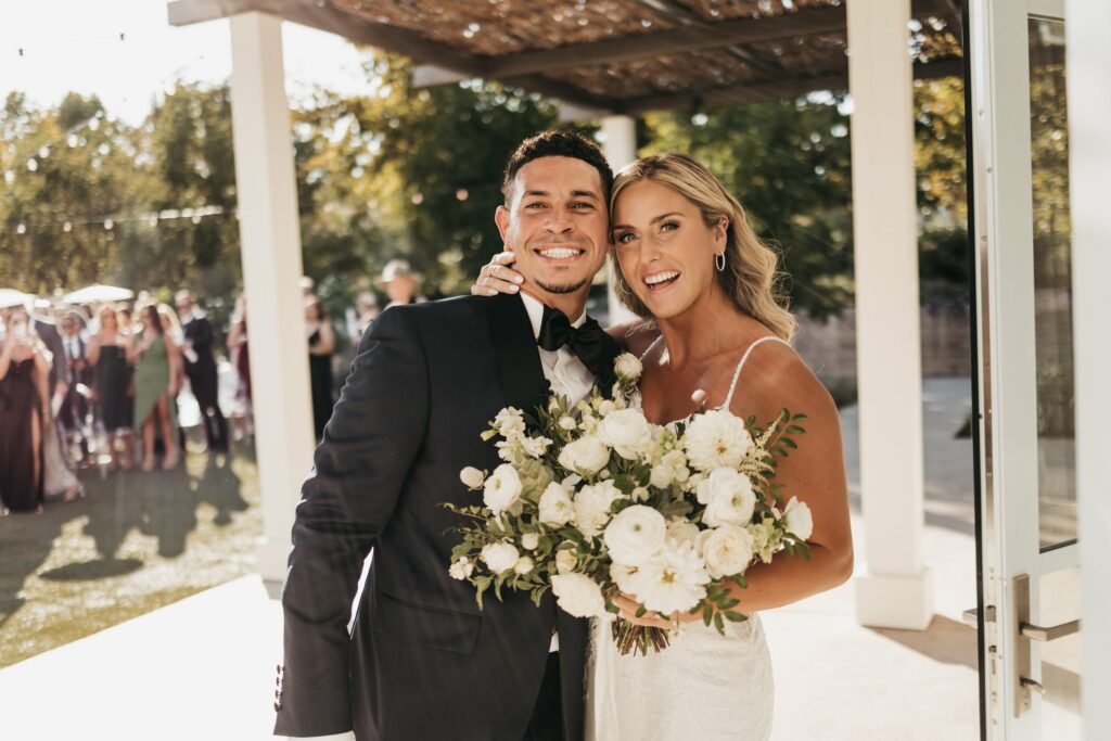 A couple dressed in wedding attire, with the groom in a black tuxedo and the bride holding a bouquet of white flowers. They are smiling and embracing, with guests blurred in the background.