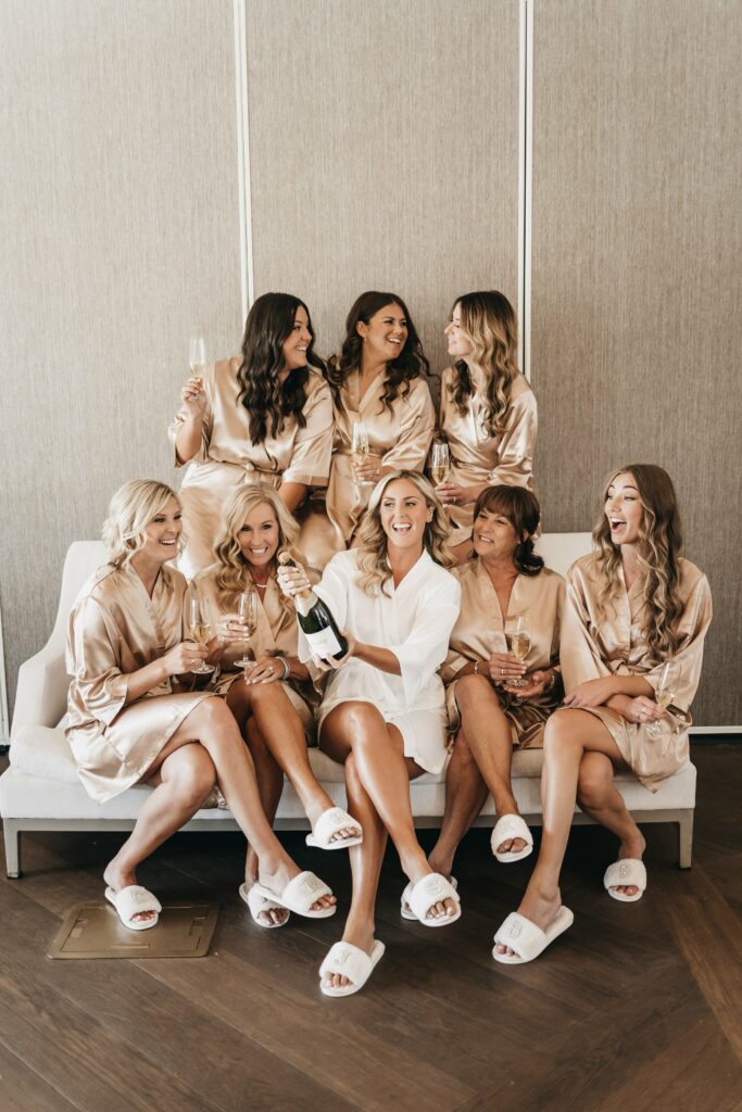 Seven women in robes, sandals, and holding glasses of wine sit on a sofa and smile together. One woman in the center holds a bottle of champagne.
