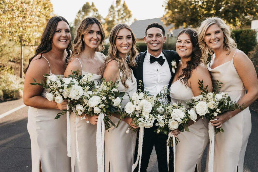 A group of six people dressed in formal attire stands together outdoors, smiling at the camera. The women are holding white floral bouquets.
