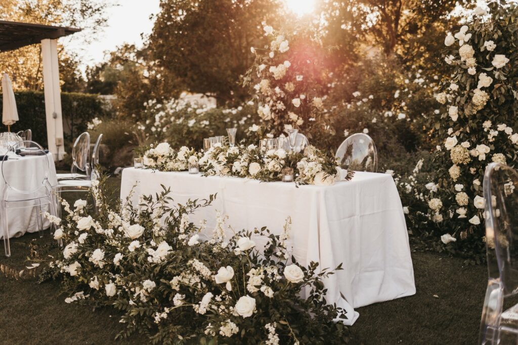 An outdoor wedding table is decorated with white flowers and greenery. Clear chairs are arranged around the table, and the scene is illuminated by sunlight.