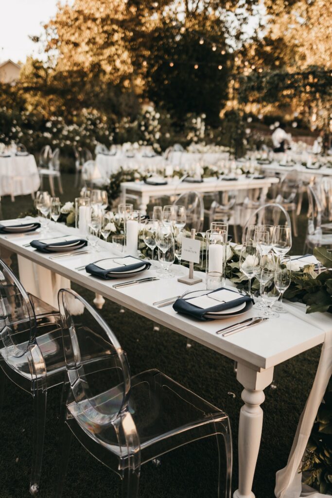A long outdoor dining table is set with plates, glassware, and candles. Transparent chairs are arranged around the table, and greenery decorates the table's centerpiece. More tables are visible in the background.