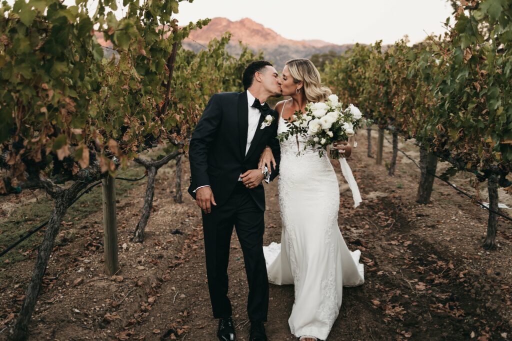 A newlywed couple kisses while walking through a vineyard. The groom is in a dark suit, and the bride is in a white gown holding a bouquet. Mountains are visible in the background.