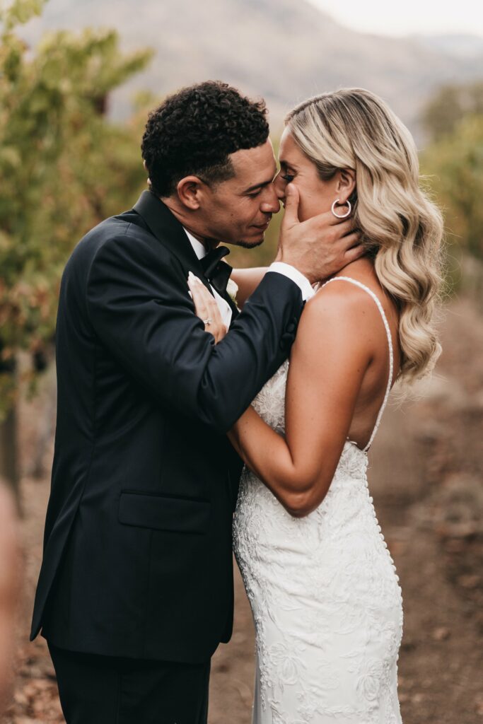 A couple dressed in wedding attire embraces outdoors, with the groom holding the bride's face closely as they share an intimate moment. The background features a nature setting with blurred greenery.