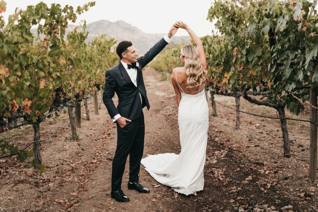 A groom in a black tuxedo twirls a bride in a white dress as they stand in a vineyard, surrounded by rows of grapevines.