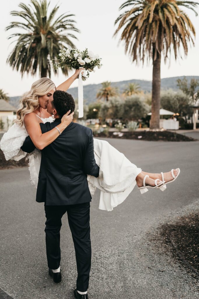 A groom in a black suit carries his bride in a white dress along a palm-tree lined street. The bride holds a bouquet above her head.
