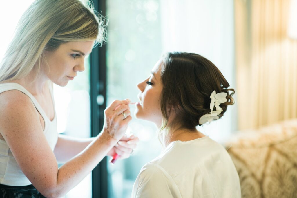 A makeup artist applies lipstick to a seated woman with her hair pinned up and adorned with white flowers in a bright indoor setting.