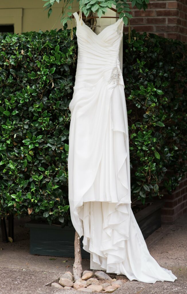 A white, strapless wedding dress with a ruched bodice and flowing skirt hangs on a wooden hanger against a backdrop of green foliage.