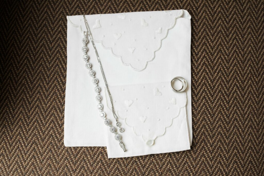A silver bracelet, necklace, and two rings are placed on white cloths with scalloped, heart-patterned edges, all atop a brown herringbone-patterned background.