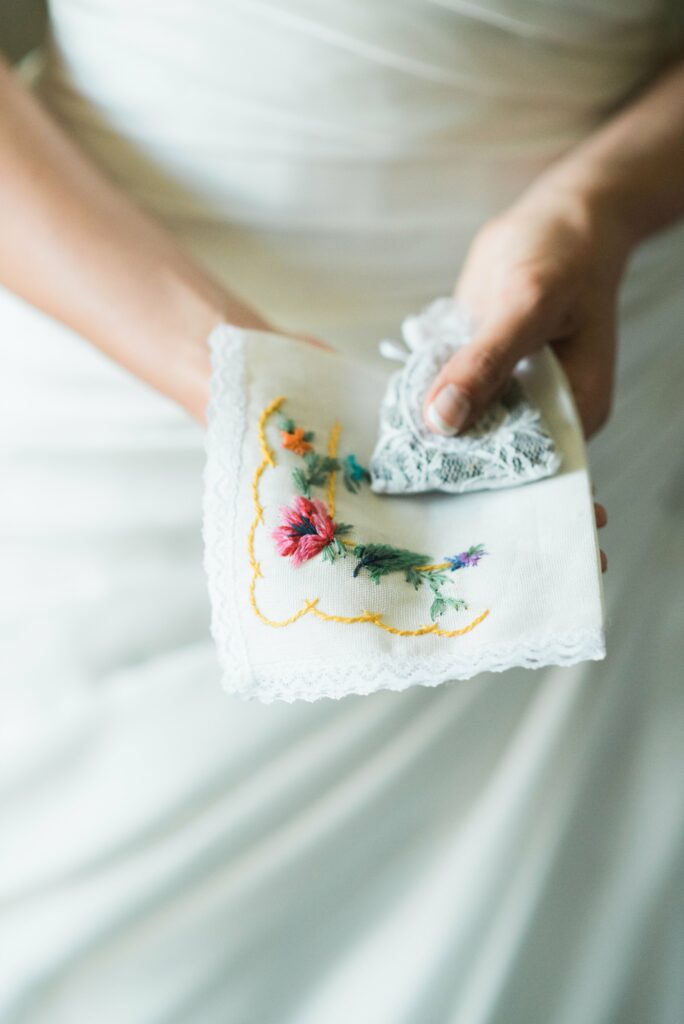 A person holds an embroidered handkerchief with floral design and lace trim, dressed in white fabric.