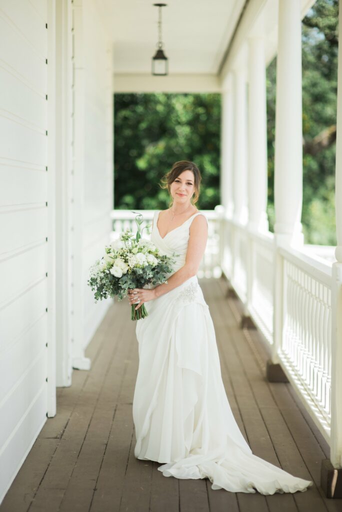 A bride in a white dress stands on a wooden porch, holding a floral bouquet, with a white railing and columns in the background.