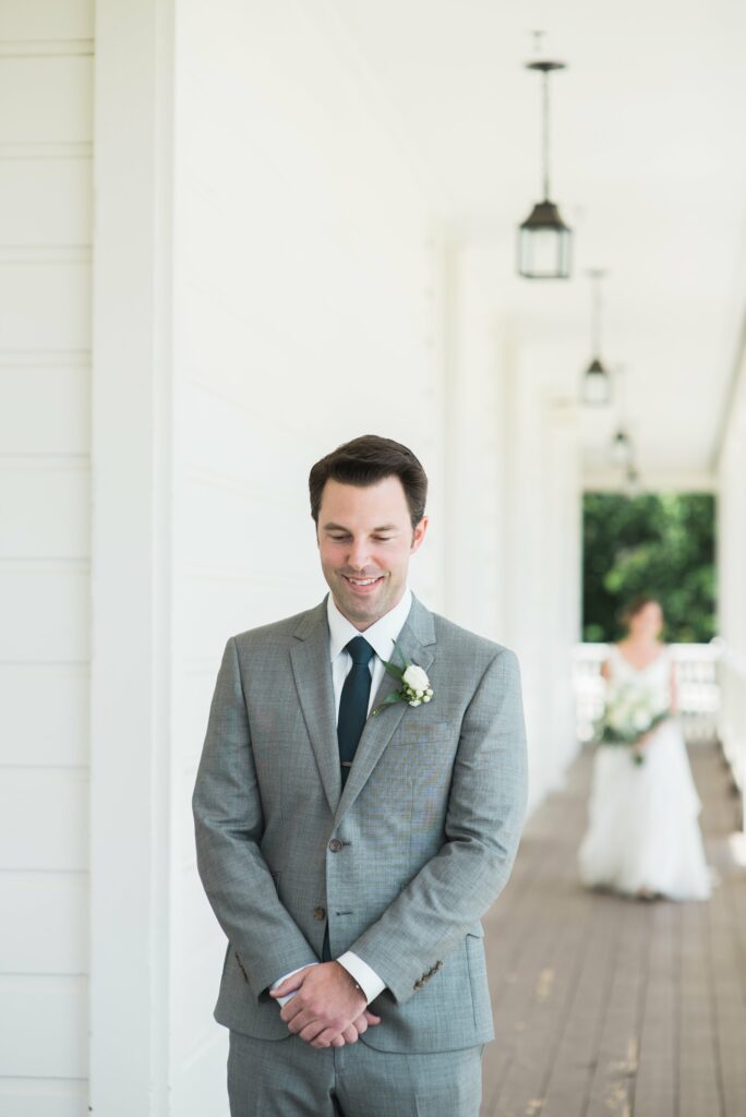 A man in a grey suit with a white boutonniere stands on a porch with his eyes closed. In the background, a bride in a white dress approaches him.
