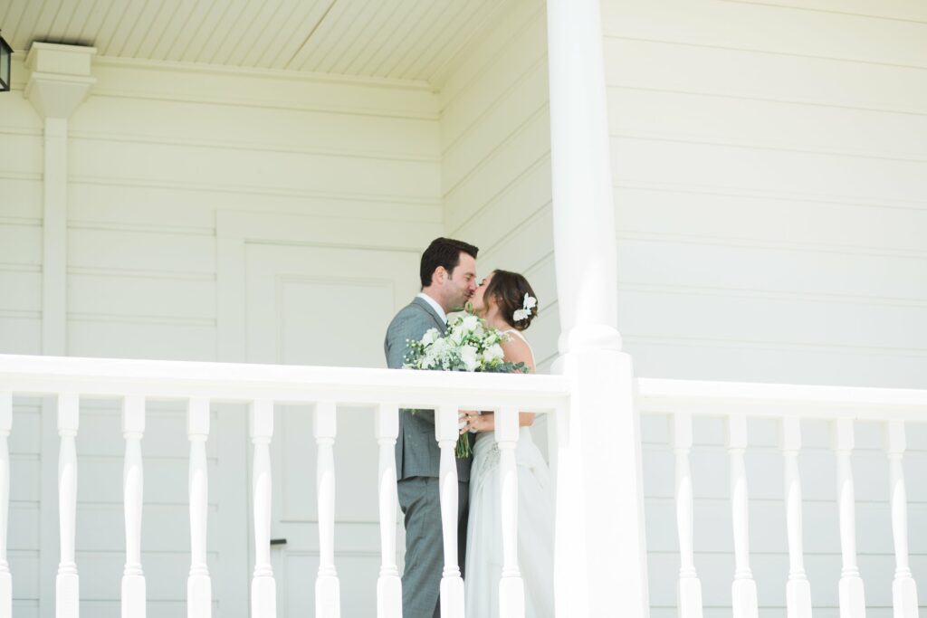 A couple dressed in wedding attire shares a kiss on the porch of a white building. The bride holds a bouquet of flowers.