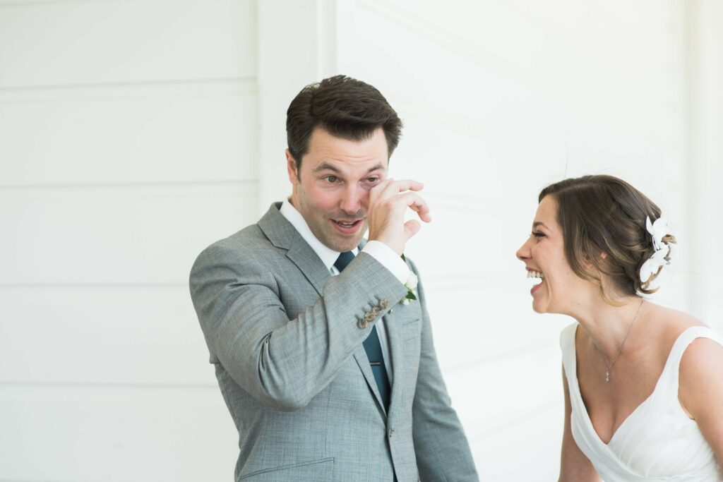 A man in a grey suit wipes a tear while a woman in a white dress smiles and laughs beside him against a white wall background.