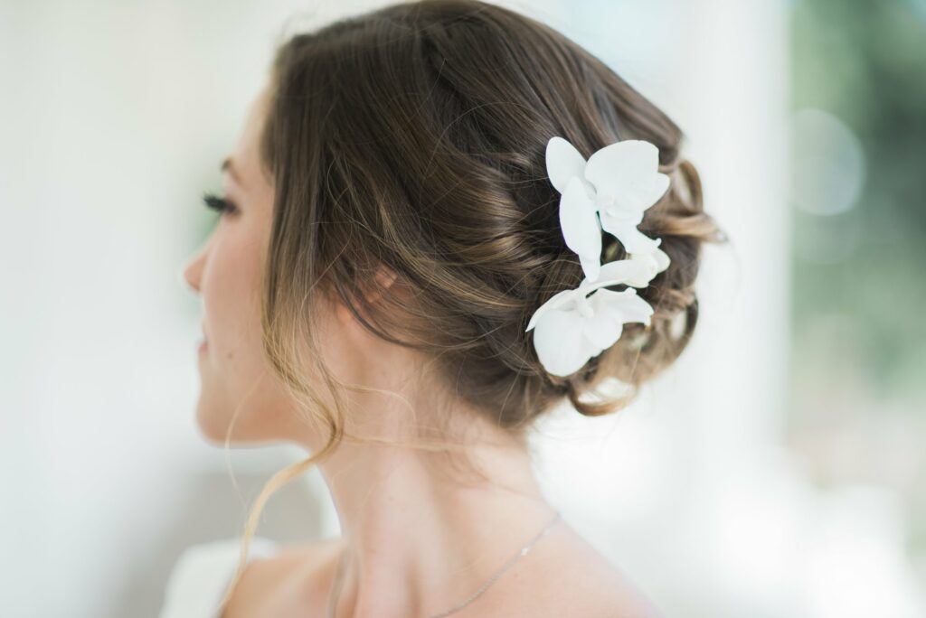 A woman with her hair styled in an elegant updo adorned with white flowers, shown from the side against a blurred background.