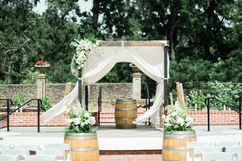 Outdoor wedding altar with a wooden canopy draped with white fabric and adorned with flowers, set on a brick platform. Two wine barrels with floral arrangements are in the foreground.