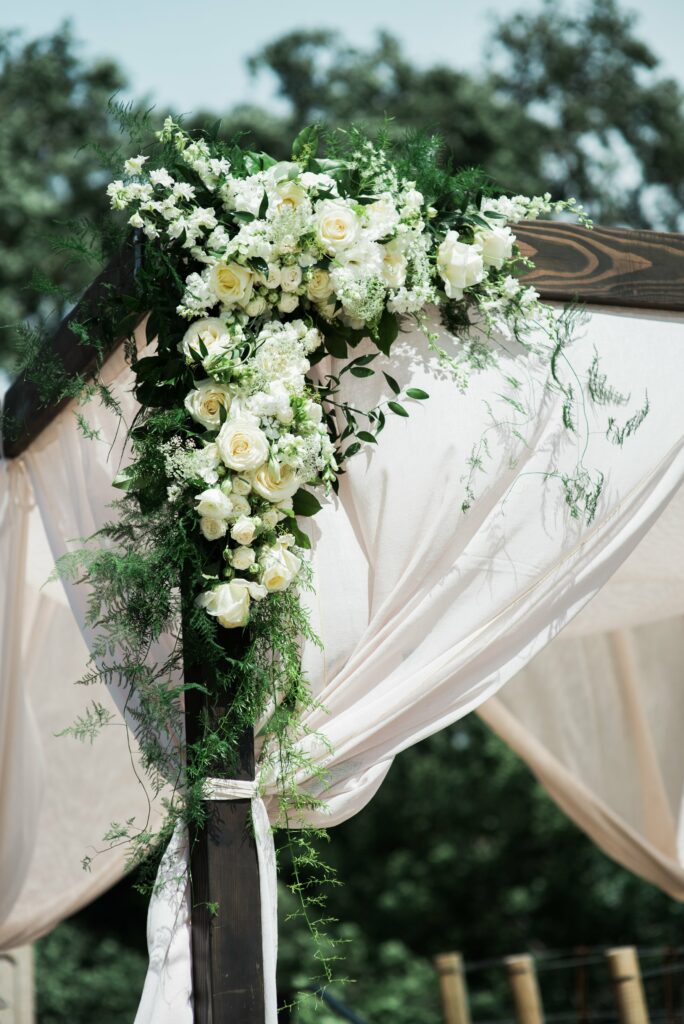 A wedding arch decorated with white roses, white flowers, and greenery under sheer white drapes on a wooden frame. Trees are visible in the background.