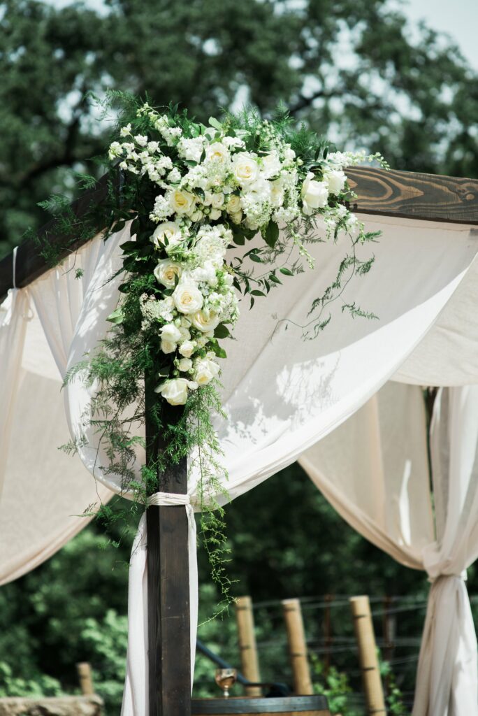 A wooden structure draped with white fabric is decorated with an arrangement of white flowers and greenery at the corner, set outdoors with trees in the background.