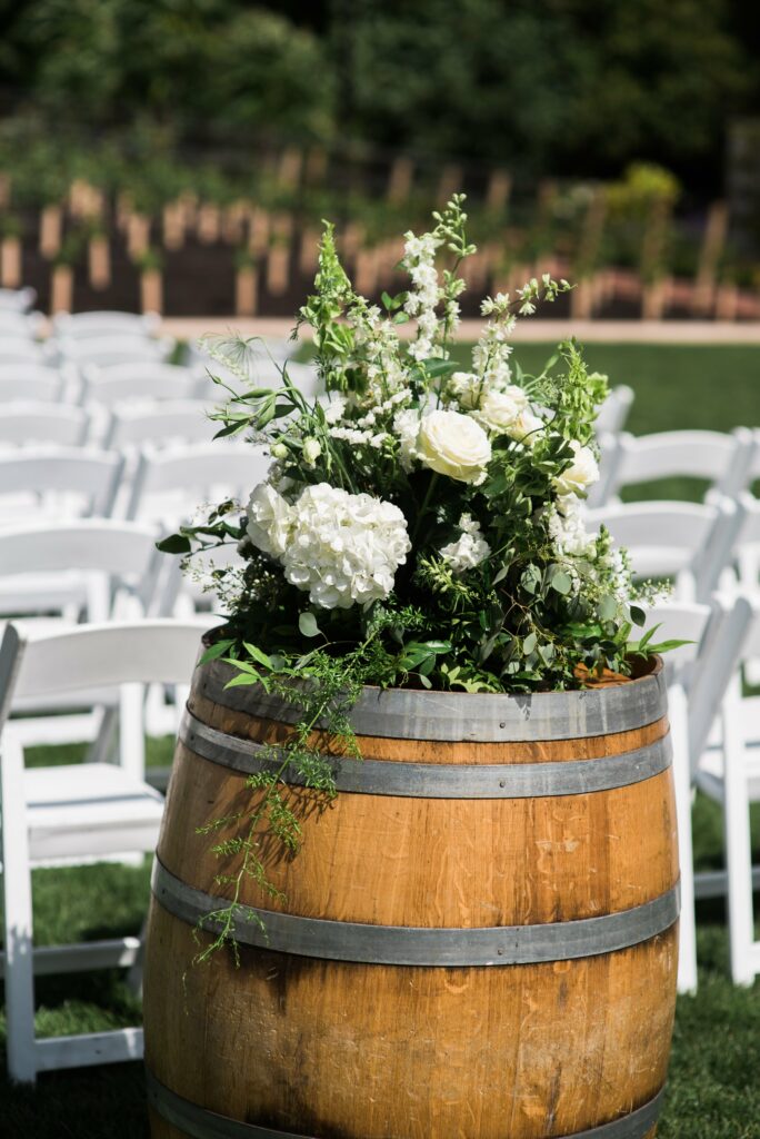 A wooden barrel adorned with white flowers and greenery stands in front of rows of white chairs set up outdoors.