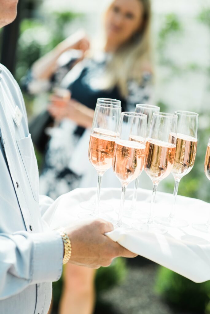 A person holding a tray with multiple glasses of rose champagne; a blurred person in the background holding a drink.