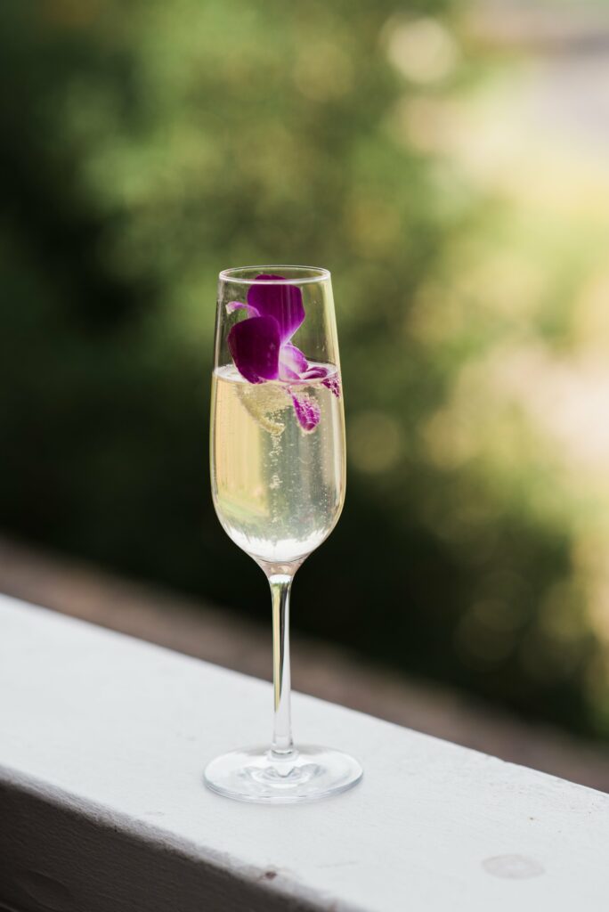 A champagne flute filled with a sparkling drink, garnished with a purple orchid, placed on a white surface with a blurred green background.
