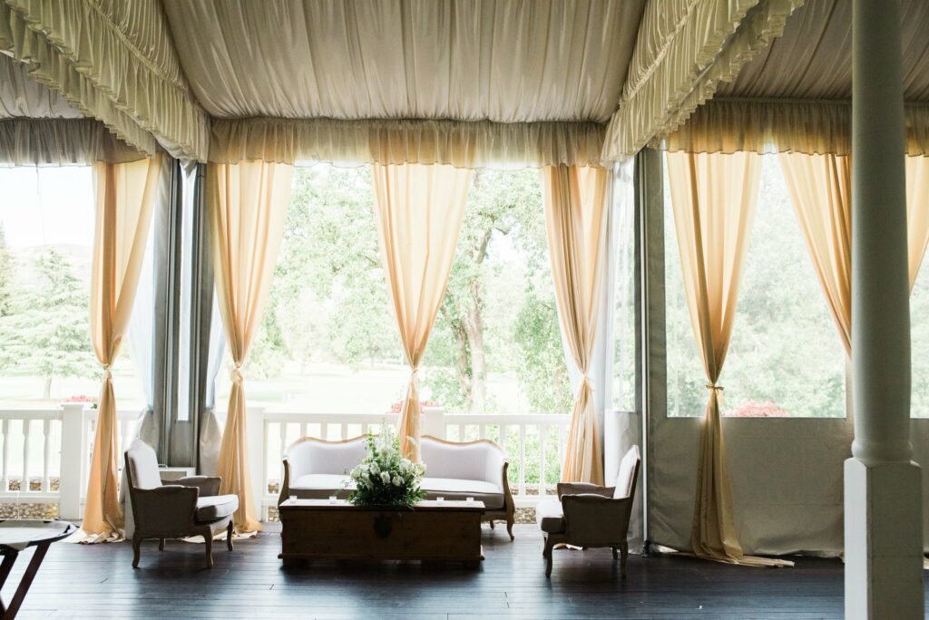 A bright, airy room with a canopy ceiling, light fabric drapes, and an elegant seating arrangement featuring two chairs and a sofa around a wooden coffee table with a flower arrangement.