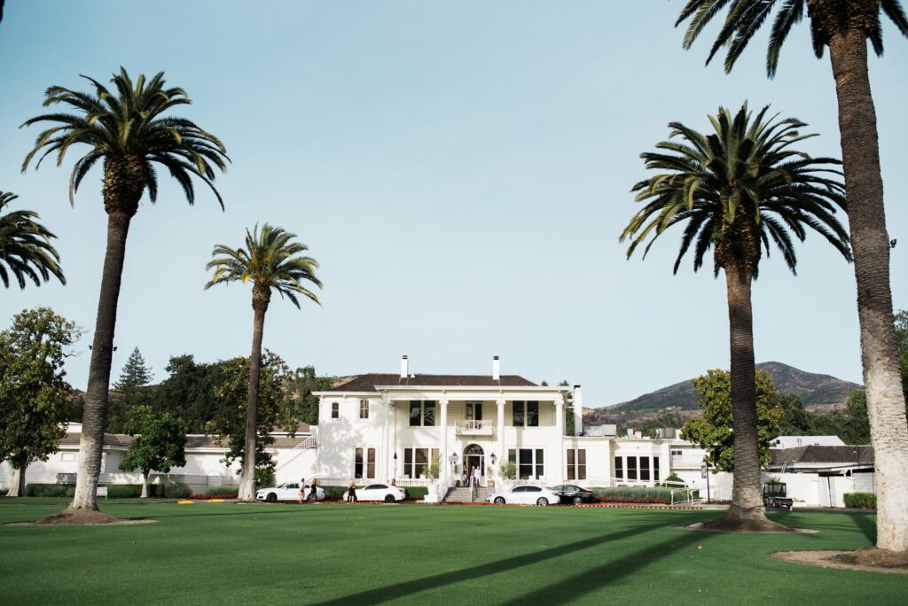 A large white building with columns, surrounded by palm trees, mountains in the background, and cars parked in front.