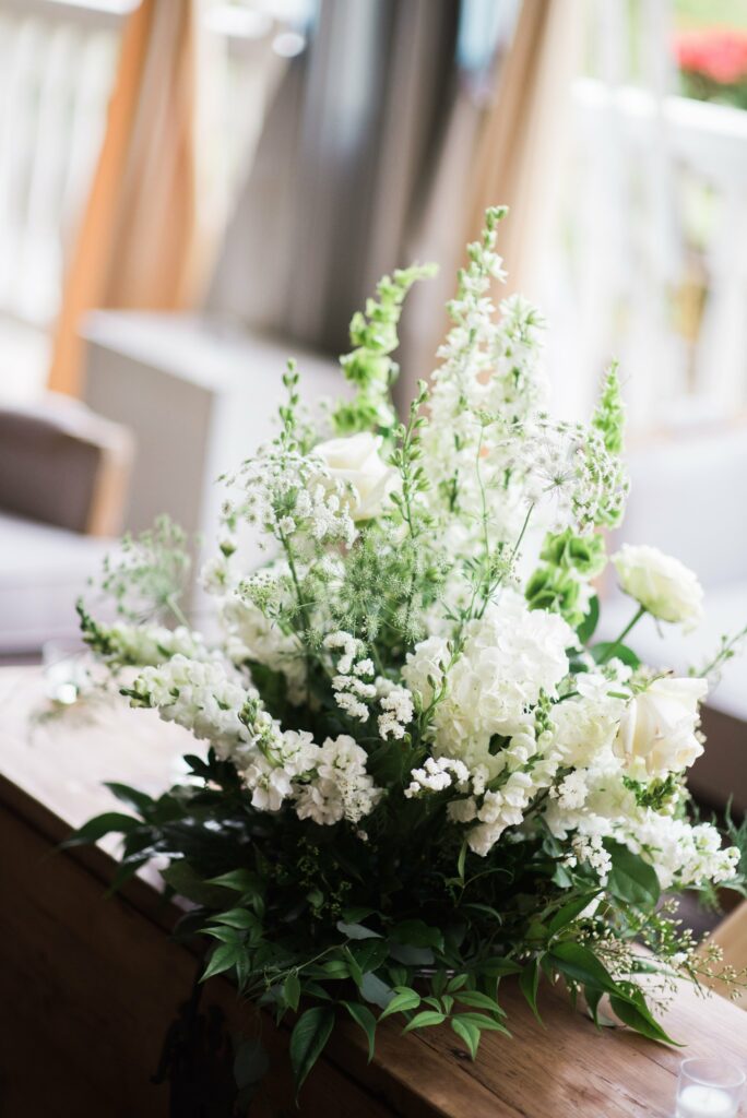 A bouquet of white flowers with green foliage arranged in a vase, placed on a wooden table with a blurred background.