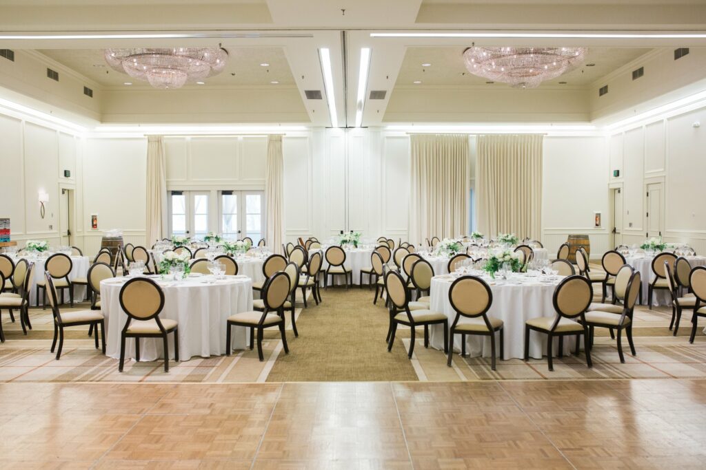 A large banquet hall with round tables set for an event. Tables are covered with white tablecloths and have floral centerpieces. Rows of beige and brown chairs surround each table. Two chandeliers hang from the ceiling.